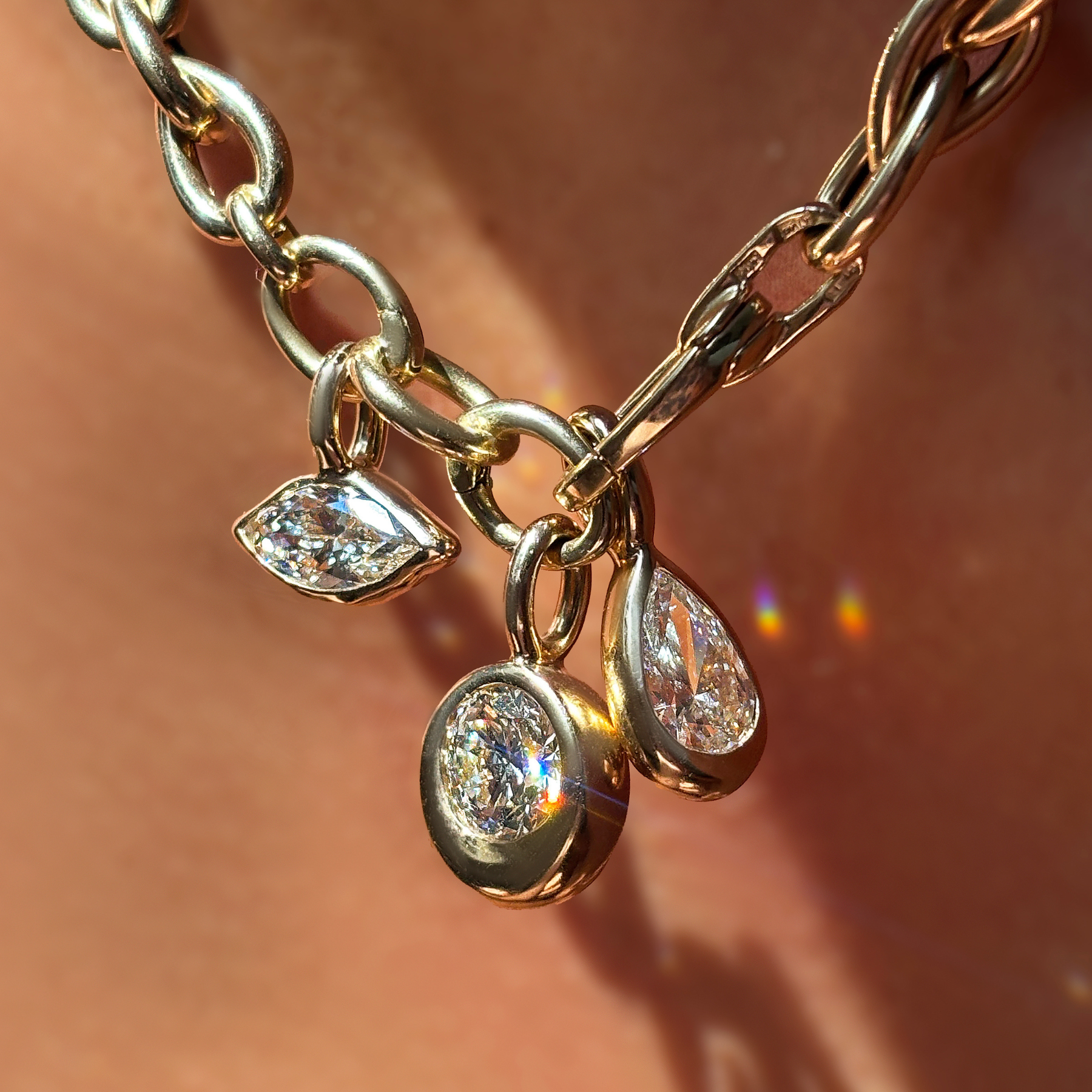 A model wears a drop link necklace in 14K yellow gold with a triple link charm lock from which hangs a marquise diamond solitaire charm, a round diamond solitaire charm, and a pear diamond solitaire charm.  All the charms are cast in 14K recycled yellow gold and an ethical lab grown diamond.
