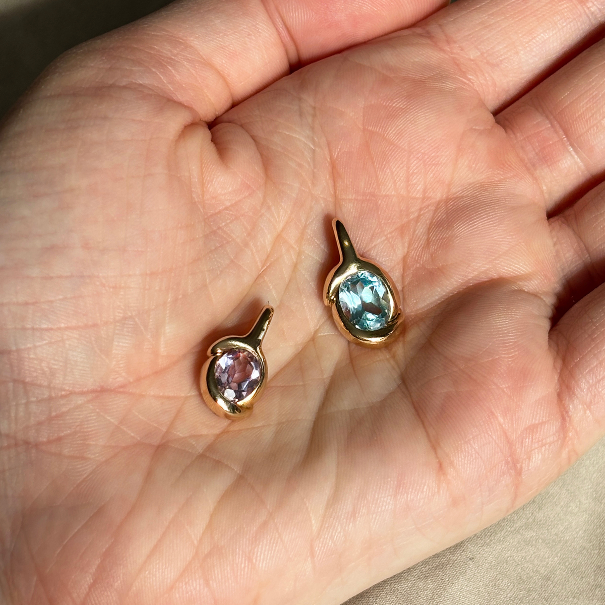 Two AMANDA PEARL molten knot charms, with blue topaz and amethyst rest in a hand, encircled in 14K recycled yellow gold bezels.