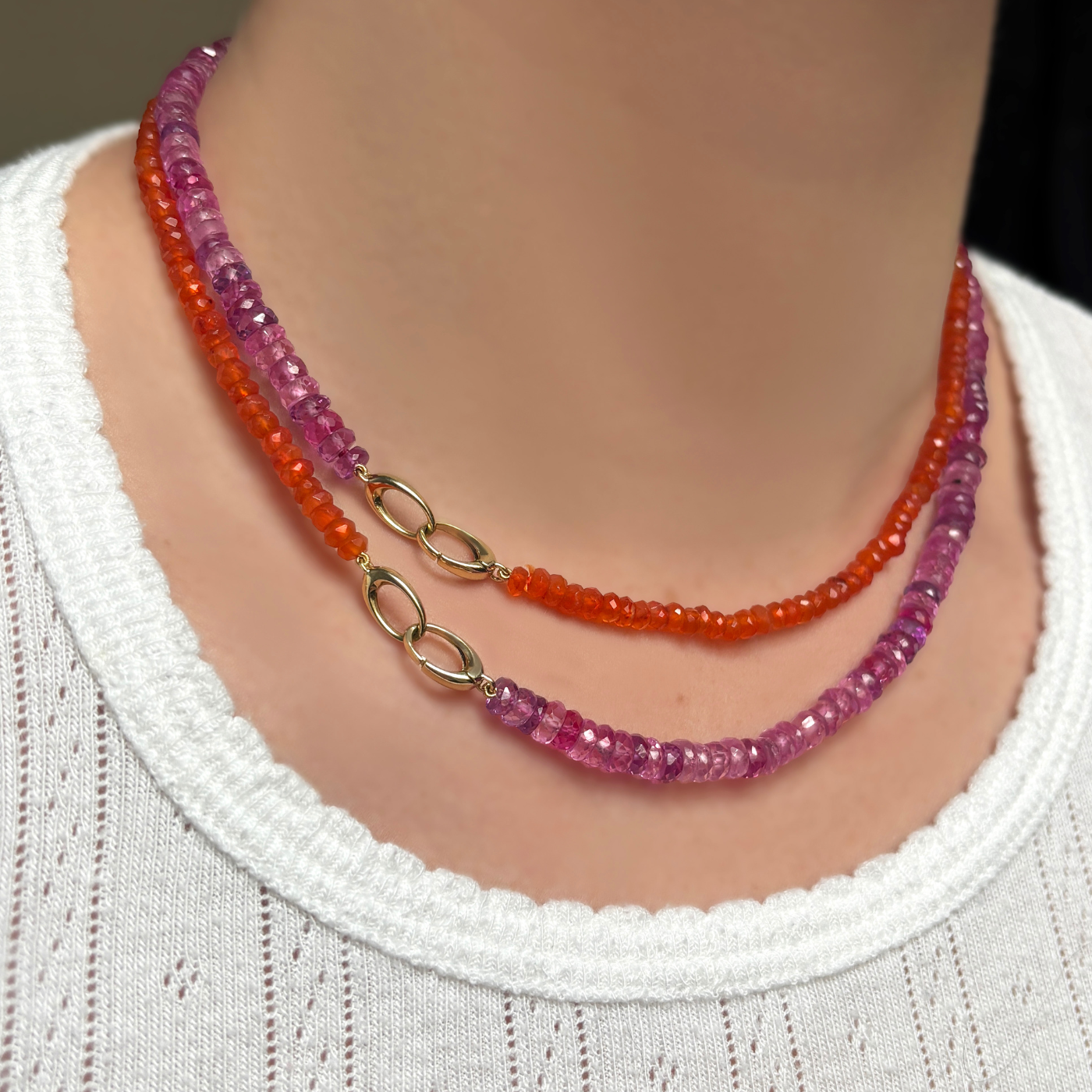 A woman in a plain white t-shirt shows wears two AMANDA PEARL necklaces. A fuchsia faceted sapphire necklace and a carnelian faceted necklace. She shows off the dramatic evolution clasp.