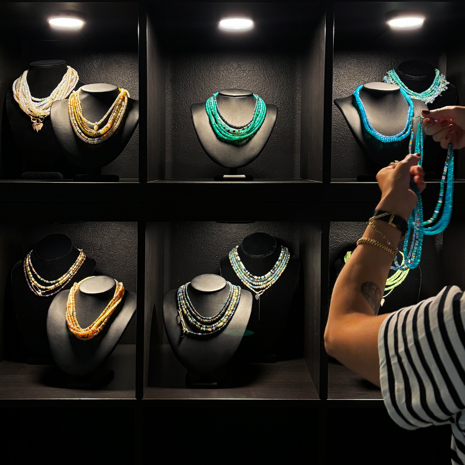Amanda Brotman's hands are shown holding a strand of blue opal beaded necklace strands, organizing them on black mannequin busts at the AMANDA PEARL showroom in NYC.