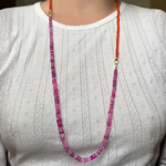 A woman in a plain white t-shirt shows how you can use your AMANDA PEARL evolution clasp to attach a fuchsia sapphire necklace to a carnelian faceted necklace to make one long, two tone necklace.
