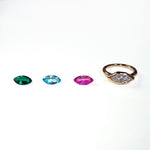 3 colorful marquise shaped gemstones next to a yellow gold molten solitaire marquise-shaped ring on a white background.