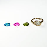 3 colorful pear shaped gemstones next to a yellow gold molten solitaire pear-shaped ring on a white background.