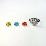 3 colorful faceted round gemstones next to a yellow gold molten solitaire round ring on a white background.