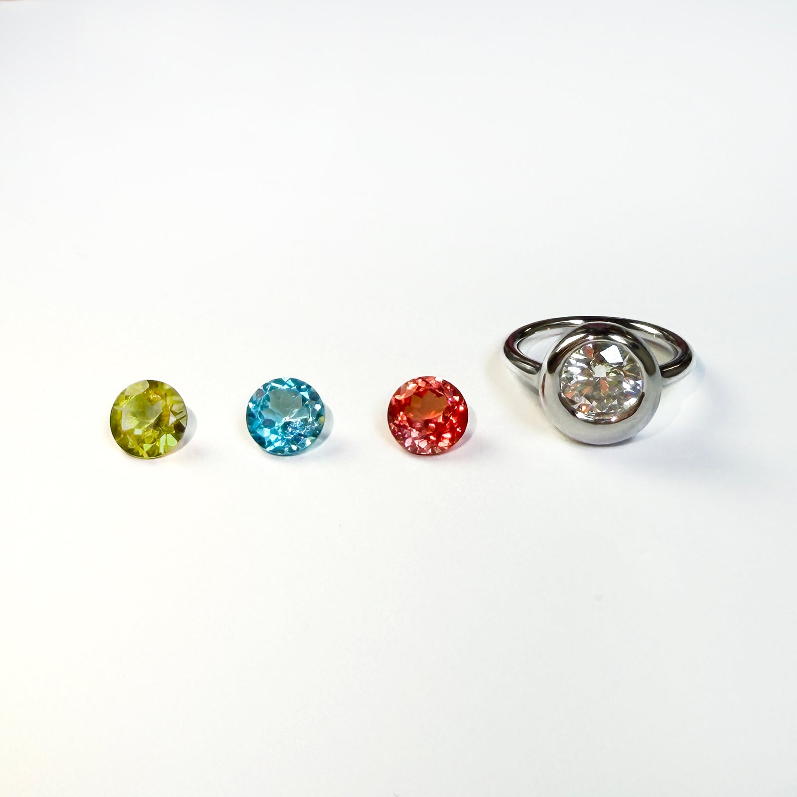 3 colorful faceted round gemstones next to a yellow gold molten solitaire round ring on a white background.