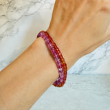 A close-up of a person's wrist wearing two beaded bracelets, one made of orange faceted padparadsha sapphire beads, one made of fuchsia faceted sapphire beads set against a light marble background.