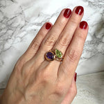 A close-up of a hand with dark red nail polish wearing two stacked gold ring set with a pear-shaped peridot gemstone and an oval amethyst gemstone, set against a white and gray marble background.