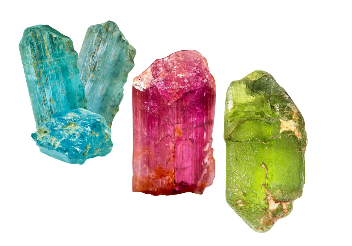 Rough uncut blue topaz, pink topaz and peridot stones sit together against a white background.
