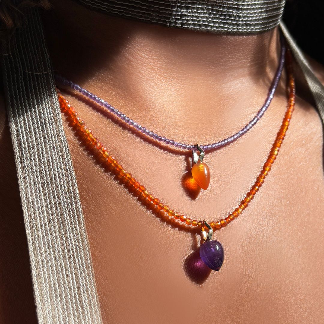 A model wears a carnelian slim gemstone necklace from AMANDA PEARL from which hangs an amethyst drop charm. She also wears an amethyst necklace necklace from which hangs a carnelian drop charm with recycled 14k ripple bales.