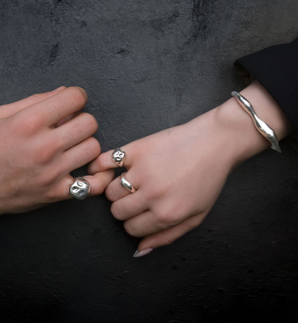 A couple locks pinkies against a black background. The man wears a skull pinky ring, as does the woman. She also wears a molten sterling ring and molten cuff bracelet, with rounded smooth lines. 