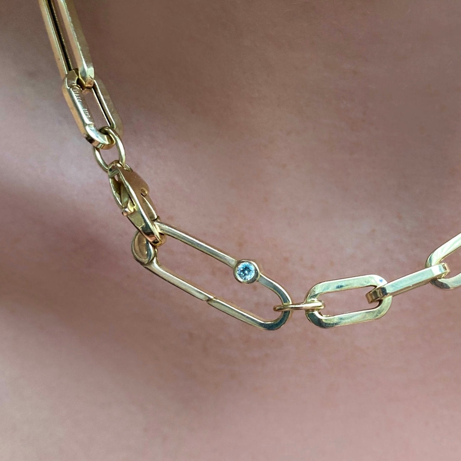 A shiny Paperclip lock charm featuring a 3mm diamond stone clasps an irregular paper clip chain around a model's neck. Both items are cast in 14k yellow gold.