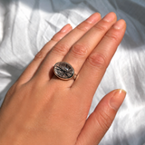 A model wears a recycled sterling silver rutilated quartz cabochon pebble ring on her hand over a white duvet cover.