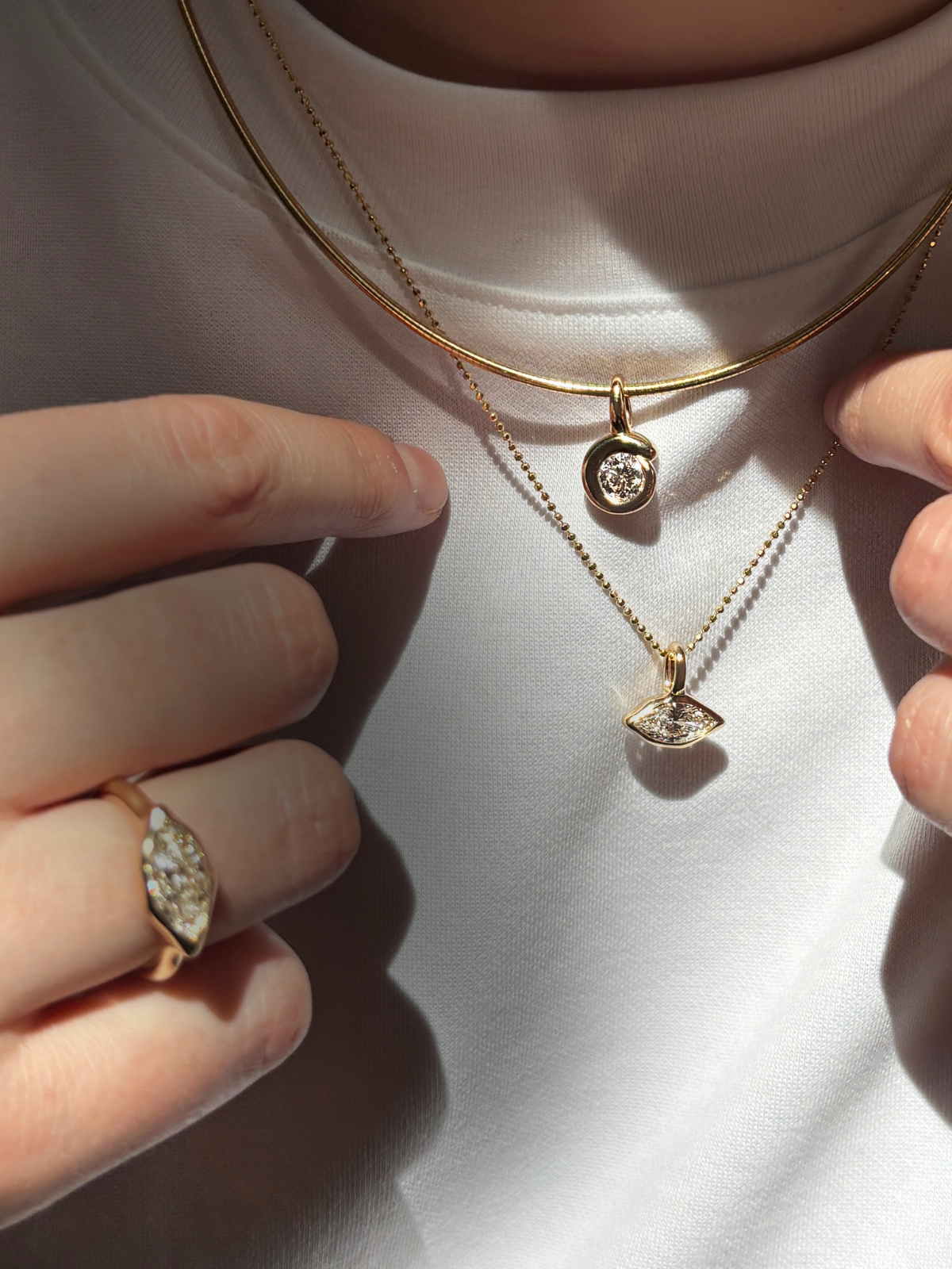 A model wears 14K recycled yellow gold charms and chains from AMANDA PEARL. A quill bezel charm with lab grown diamond on an omega chocker. A marquise diamond charm won a diamond cut bead chain, and a marquise diamond ring.