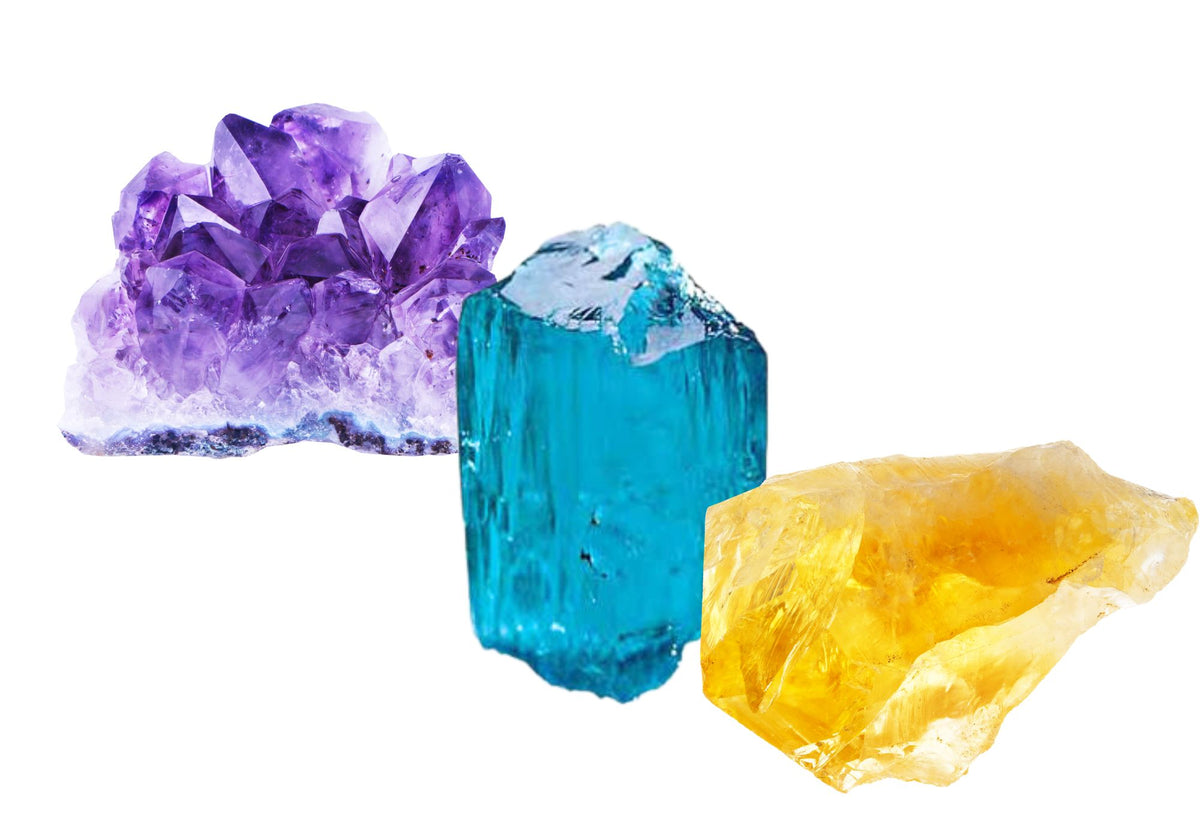 Rough, uncut amethyst, paraiba toumaline and citrine crystals sit grouped together on a white background. 