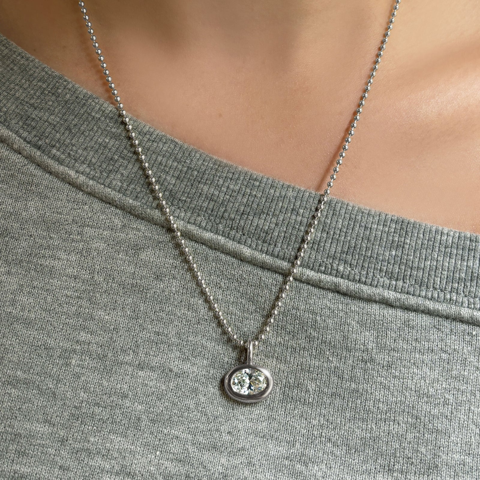 Molten Solitaire Charm in white gold with an oval diamond, on a white gold bead chain worn over a heather grey sweatshirt.