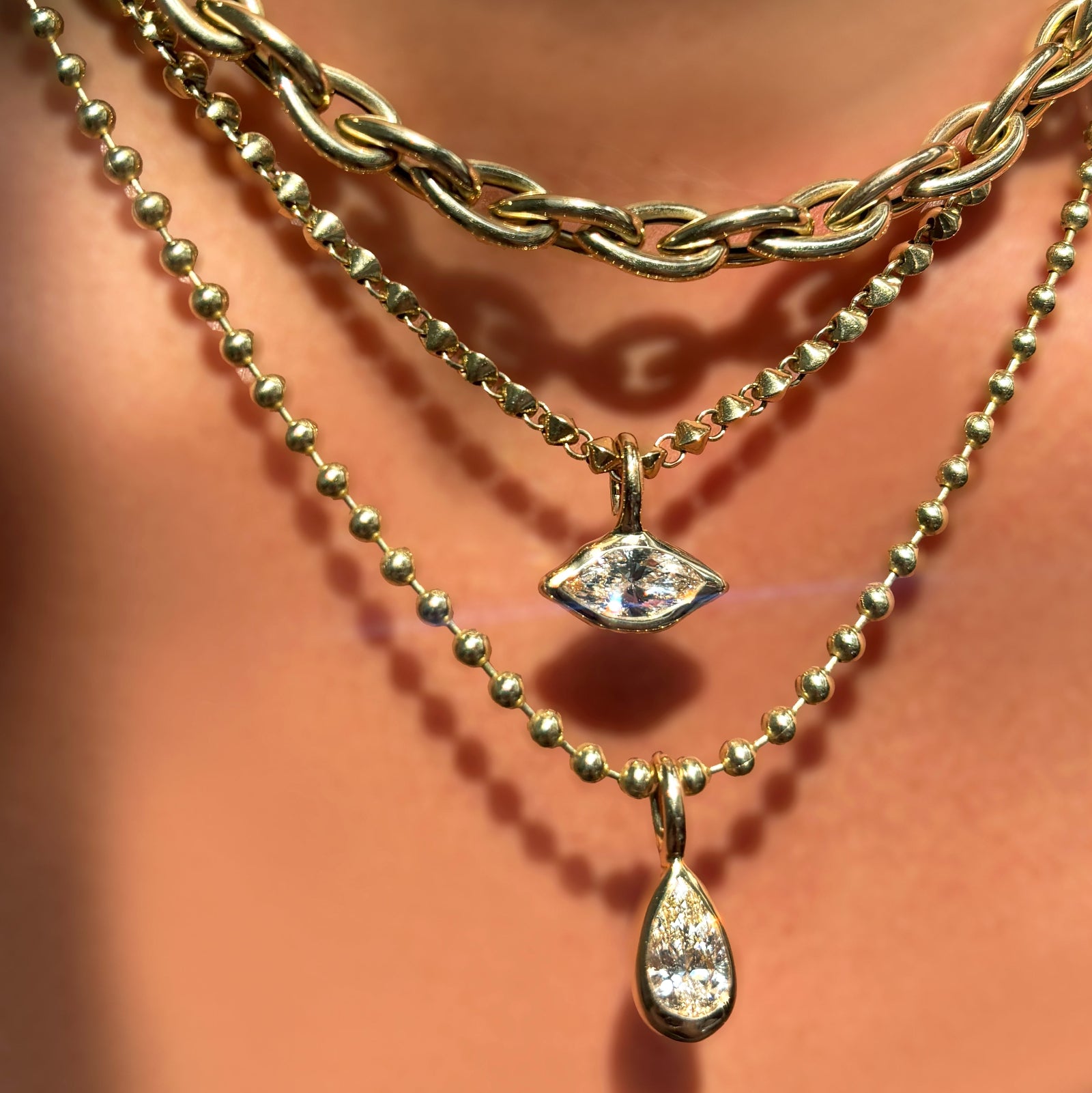 Drop link chain necklace layered over a tibet chain with molten solitaire charm with marquise diamond layered over  a 3mm bead chain with molten solitaire charm with pear cut diamond on a model's neck.