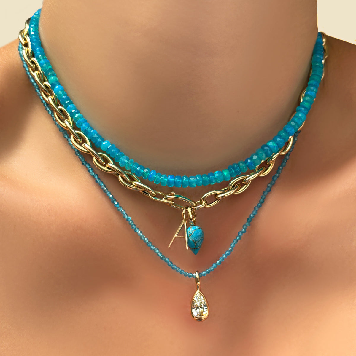 bright blue faceted opal necklace with  drop chain necklace, letter A charm, turquoise drop charm, blue apatite slim gemstone necklace, and molten pear solitaire charm on a model's neck.