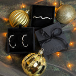 A holiday assemblage of gold ornaments, fairy lights, and black gift boxes, holding white gold pavé ripple earrings and a full pavé ripple bracelet. 