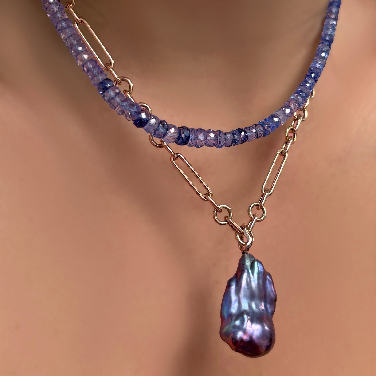The AMANDA PEARL denim blue faceted sapphire necklace layered over an irregular cable chain necklace with Storm blue baroque pearl on 14k yellow gold ripple bale on a model's neck.