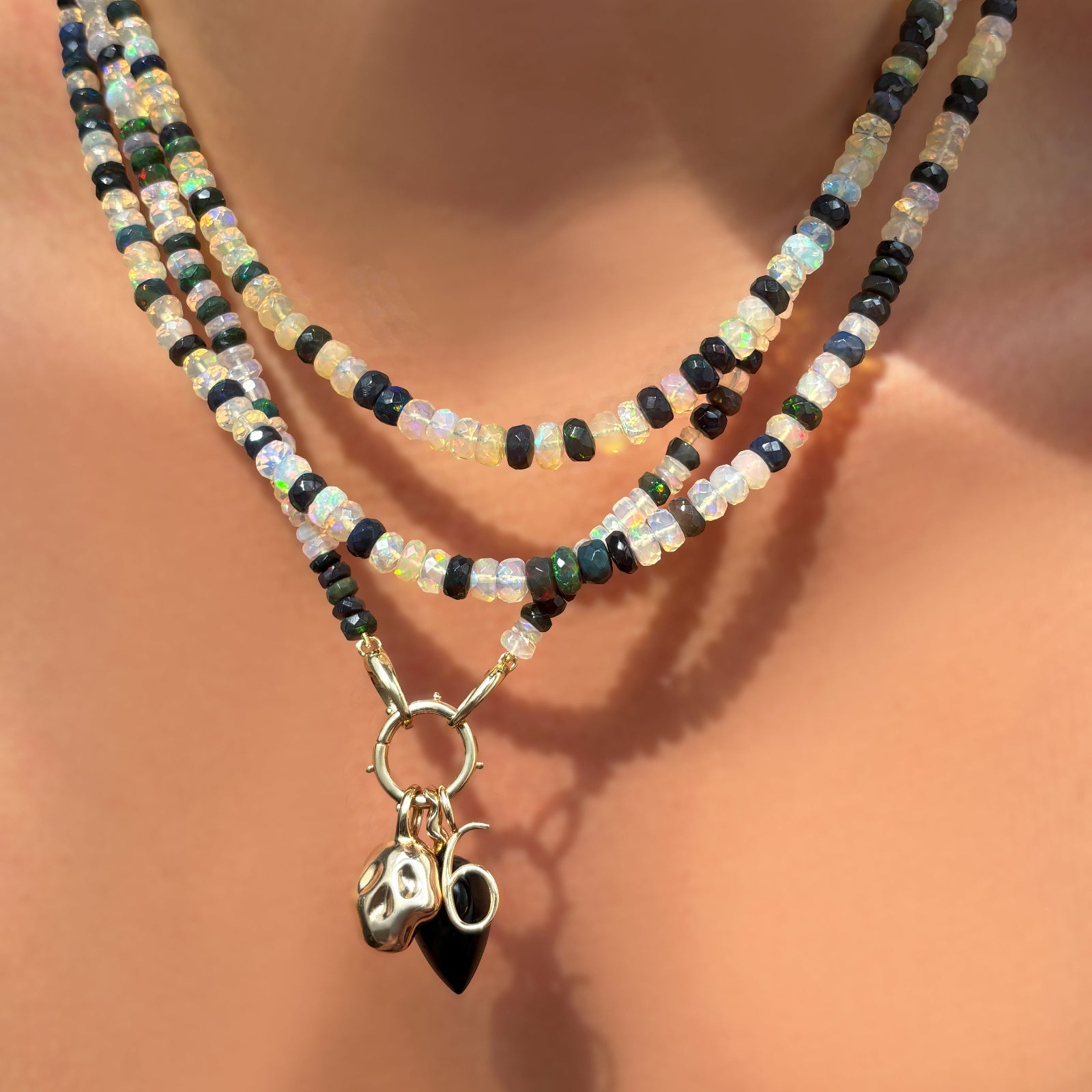 Double shade welo faceted opal necklace with beaded round charm lock dropping a molten skull charm, number 6 charm, and black agate drop charm on a model's neck.