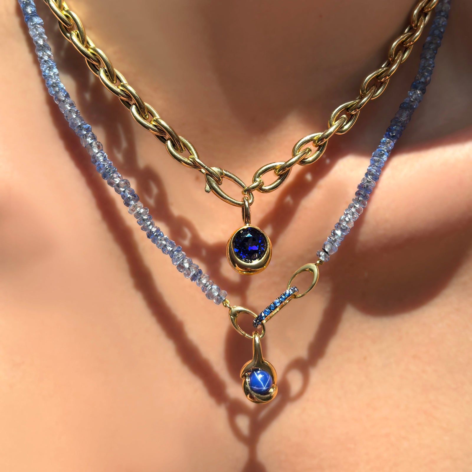 Drop link chain necklace with molten solitaire charm with round sapphire, layered with denim blue faceted sapphire necklace with small sapphire charm lock and molten knot charm with star sapphire on a model's neck.