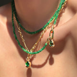 Faceted sapphire necklaces layered with irregular cable chan holding a molten solitaire charm with emerald, molten knot charm with rose cut emerald, and small emerald charm lock on a model's neck.