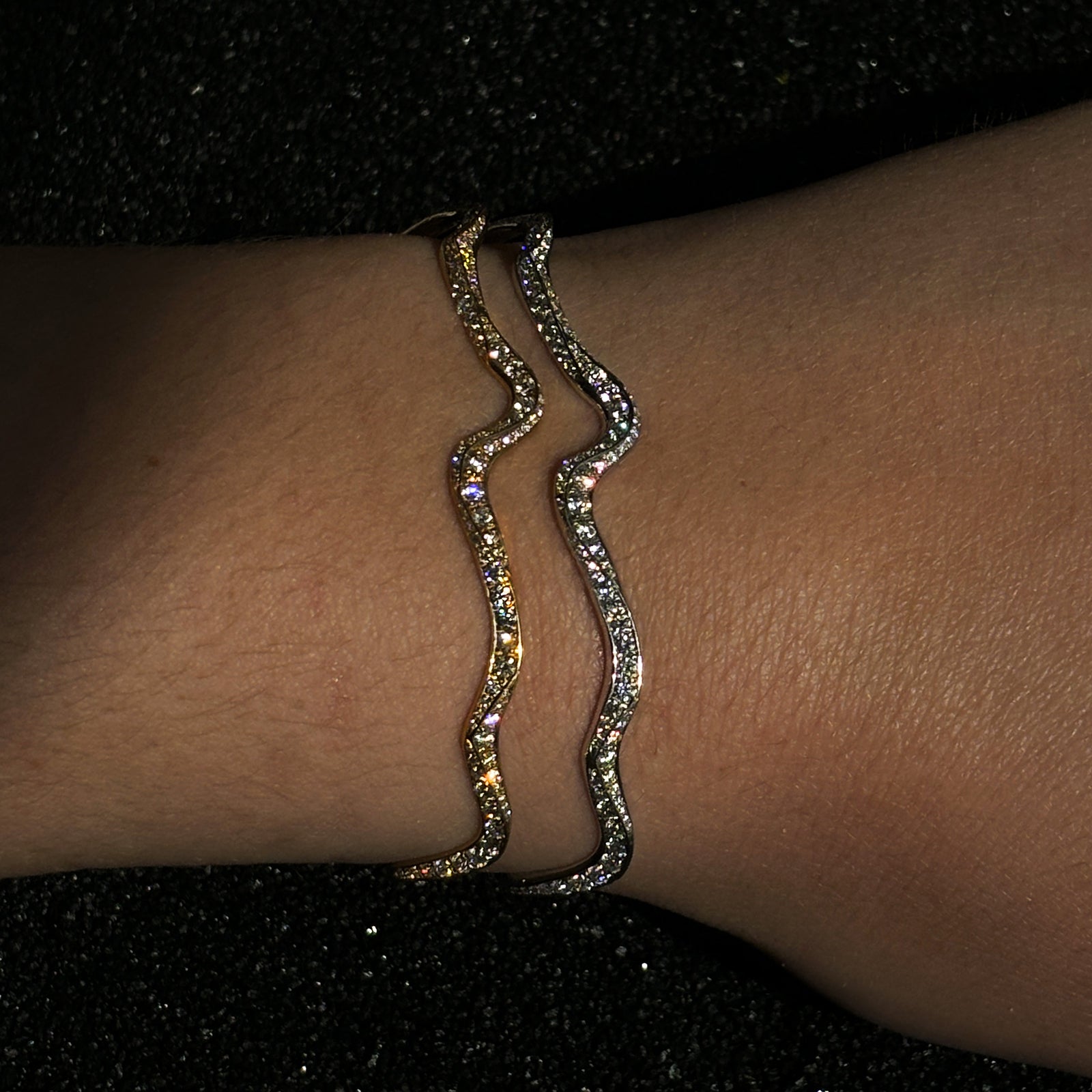 A model in dim low lighting wears a A 14K white gold, and a 14K yellow gold full pavé ripple cuff bracelet . Both feature stunning lab grown diamonds. 