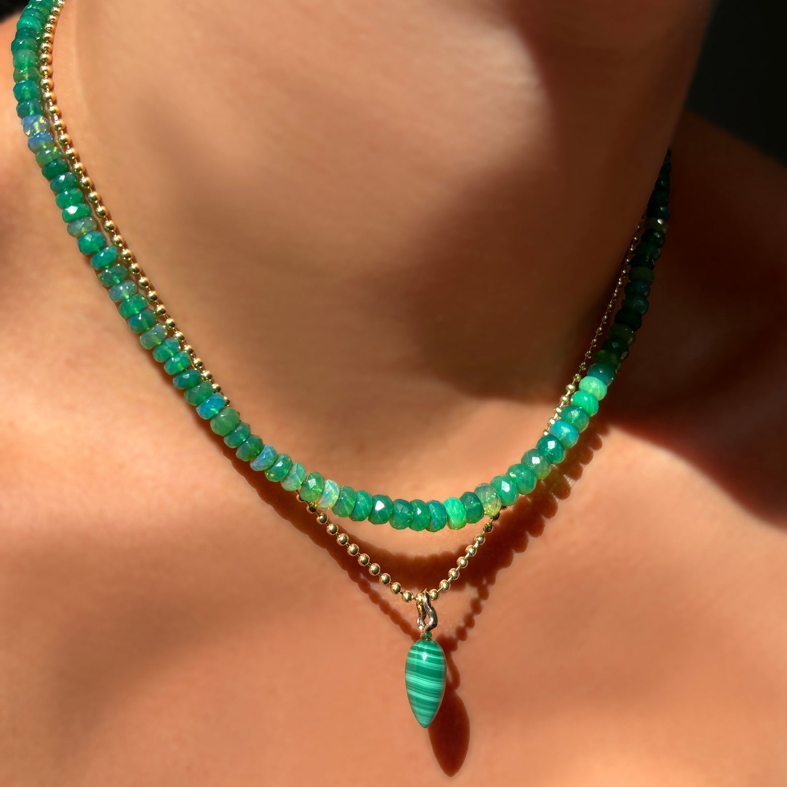 A model wears a green grass faceted opal necklace, with a 14K yellow gold bead chain, from which dangles a malachite drop charm with a 14K yellow gold ripple bail. 