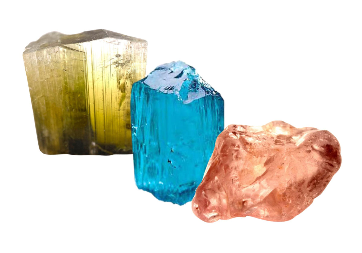 Rough uncut green tourmaline, blue topaz, and padparadscha sapphire stones sit together against a white background.