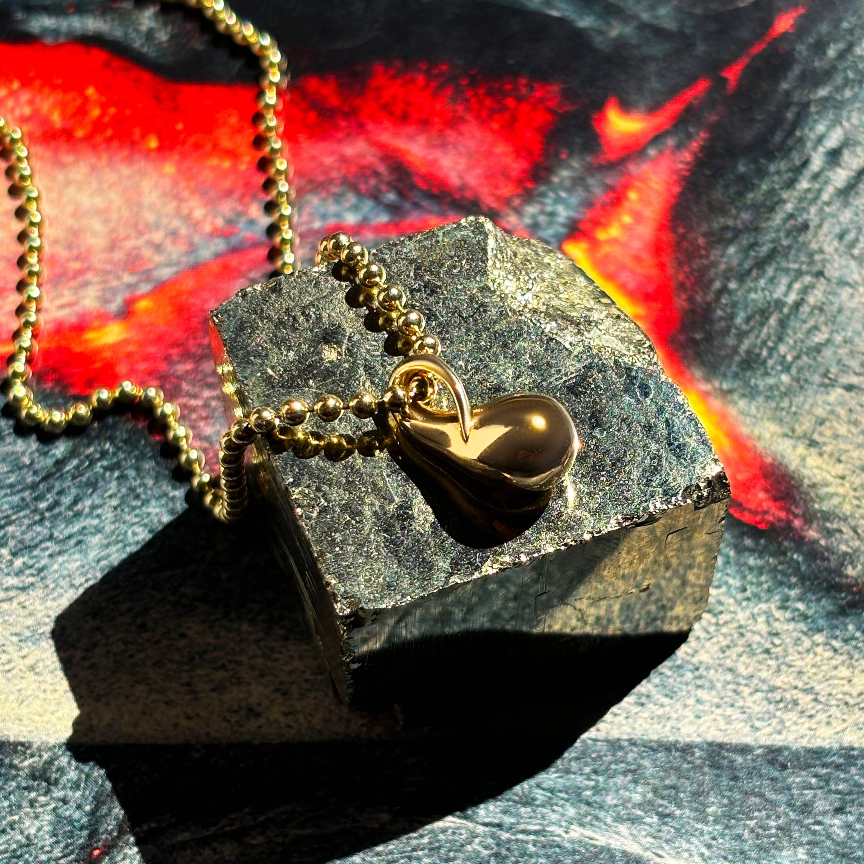 14k recycled Gold Molten Drop Charm on a gold bead chain layered over a molten lava image