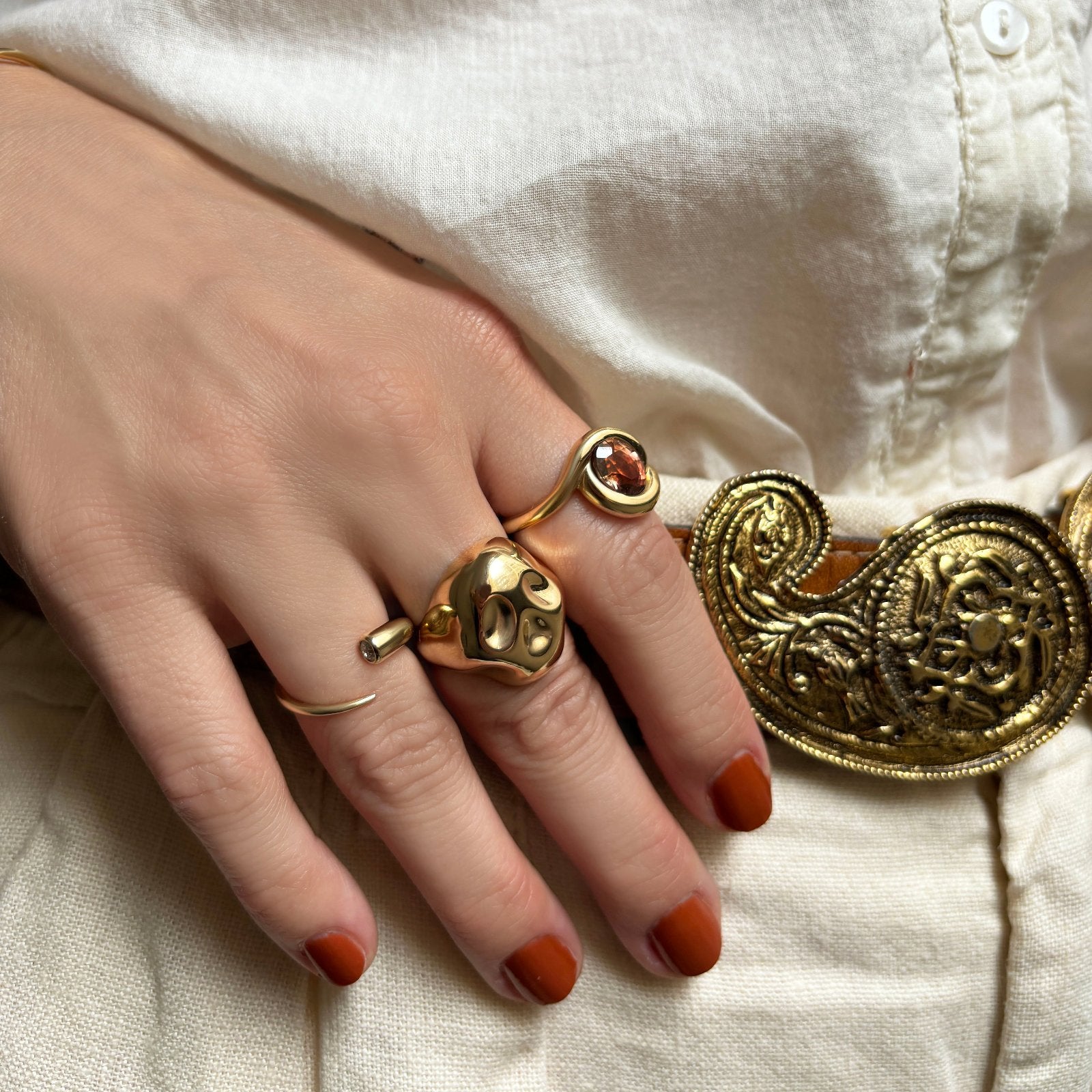 A model's hand clutches her brown belt, showing off her 14K yellow gold Quill Bypass Ring with Diamond, which is styled with the molten Skull ring and molten knot ring.
