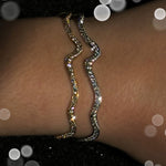 A model in a darkened space wears the 14K recycled yellow gold full pavé ripple cuff, and the white gold version. Sparkling lights cause the bracelets to glitter.