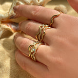 A whole handful of ripple rings set against the backdrop of beach sand and shells. The model wears 14K recycled yellow gold Ripple rings with marquise diamond stacked with Full Pavé and Plain Ripple Ring on a hand. All diamonds featured are ethical and lab grown.