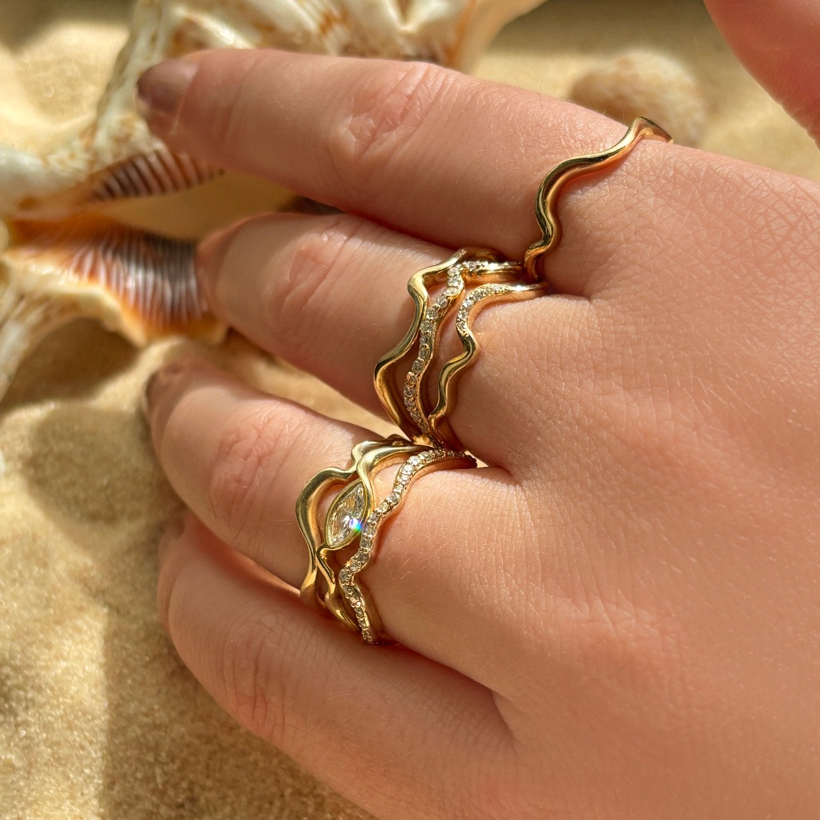 A whole handful of ripple rings set against the backdrop of beach sand and shells. The model wears 14K recycled yellow gold Ripple rings with marquise diamond stacked with Full Pavé and Plain Ripple Ring on a hand. All diamonds featured are ethical and lab grown.