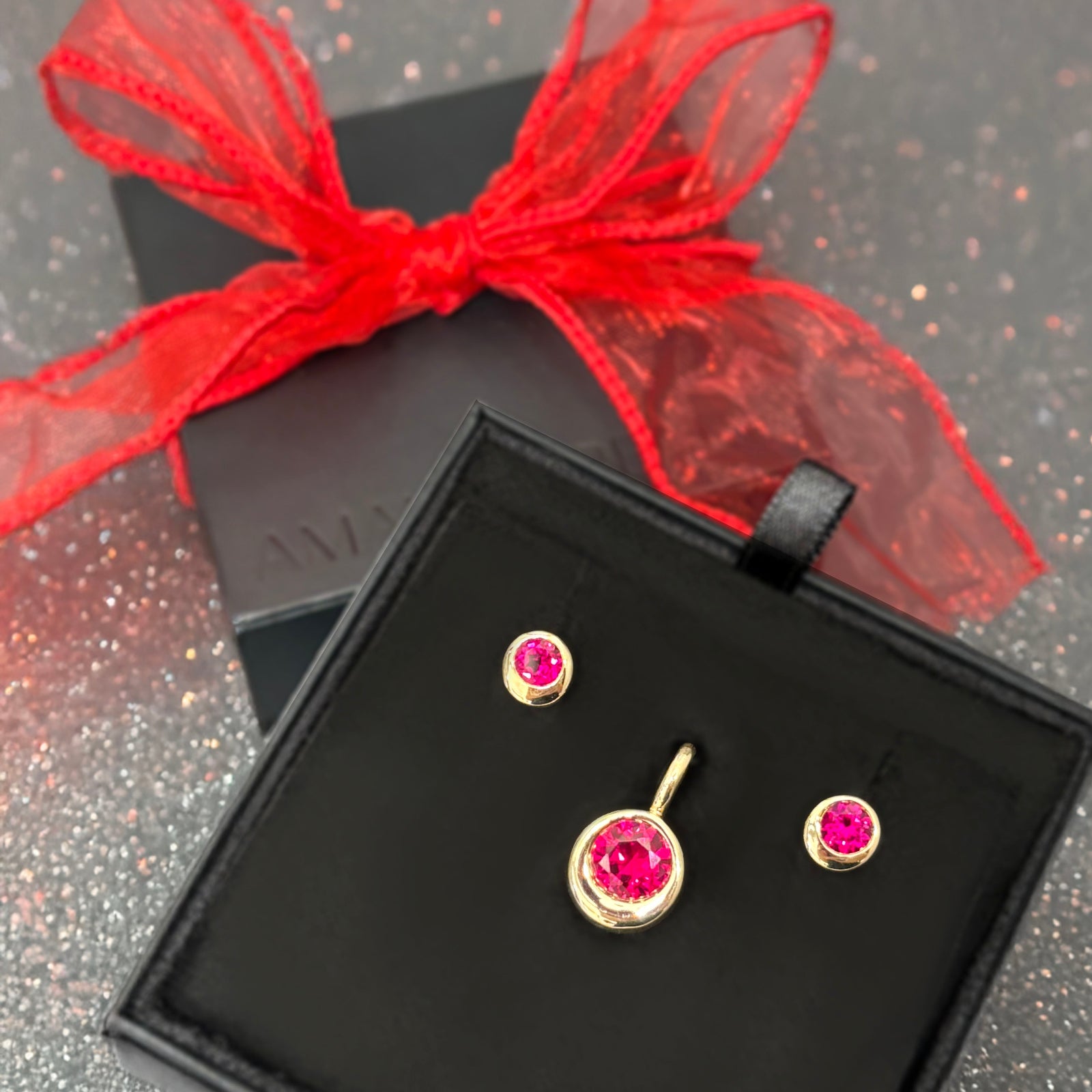 A black gift box with a pair of molten solitaire studs with round rubies and a molten solitaire charm with a round ruby.