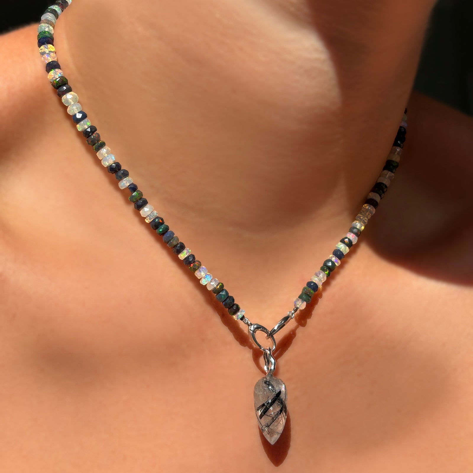 A model wears a faceted double shade welo opal necklace, with a rutilated quarts drop charm with 14K white gold ripple bail, secured to the necklace by its 14K white gold clasp. 