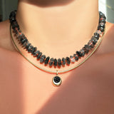 Necklace with rutilated quartz beads strung on a neon red thread layered with a gold chain and round yellow gold charm set with a black onyx stone.