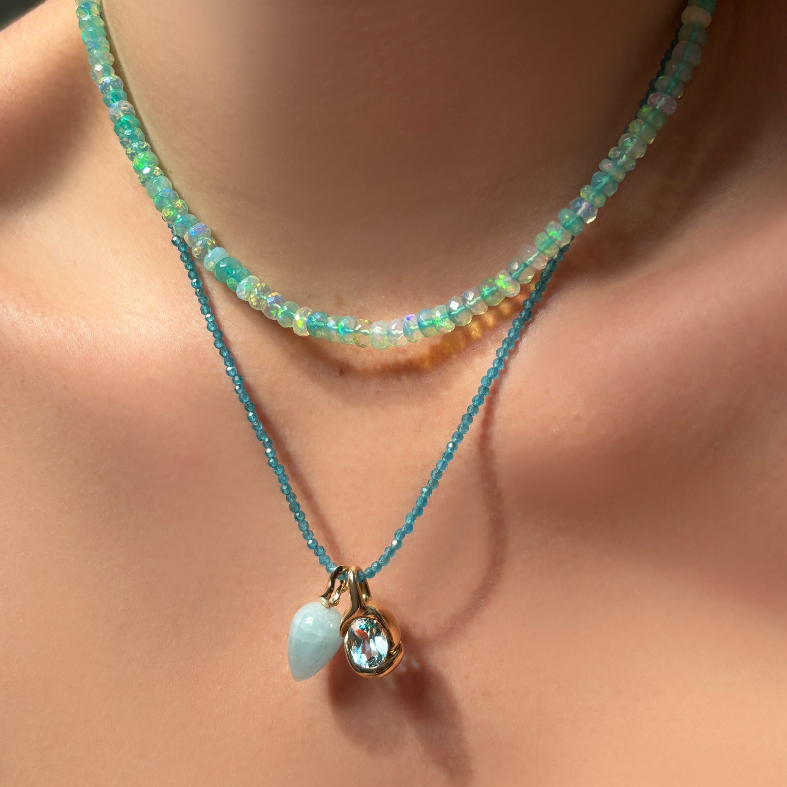 Sea glass green faceted opal necklace, layered with an apatite slim gemstone necklace with an aquamarine drop charm and molten knot charm with blue topaz hanging from it, on the body.