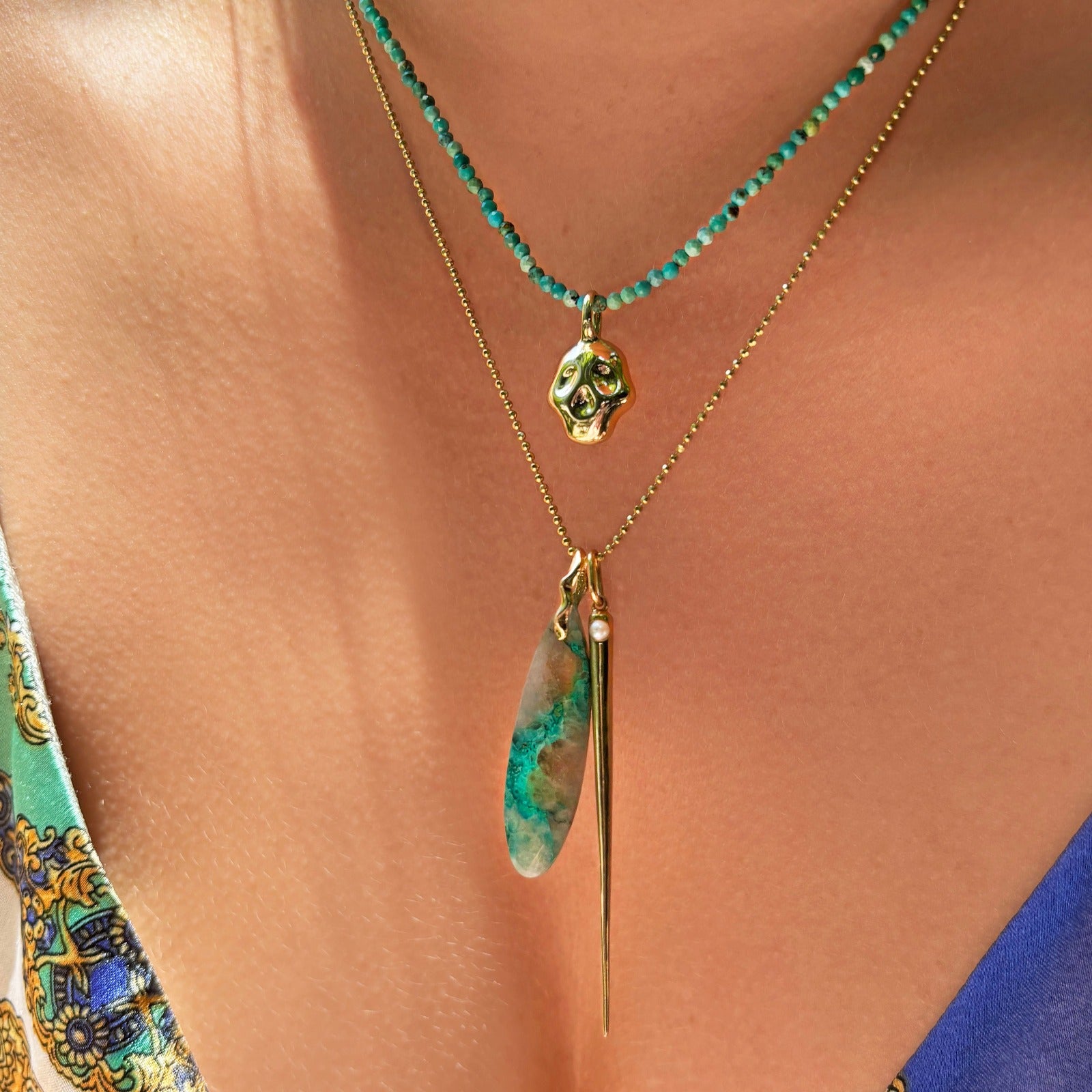 The turquoise slim gemstone necklace with a yellow gold skull charm and a yellow gold beaded necklace with a turquoise surfboard charm and a spike charm with a pearl accent. 