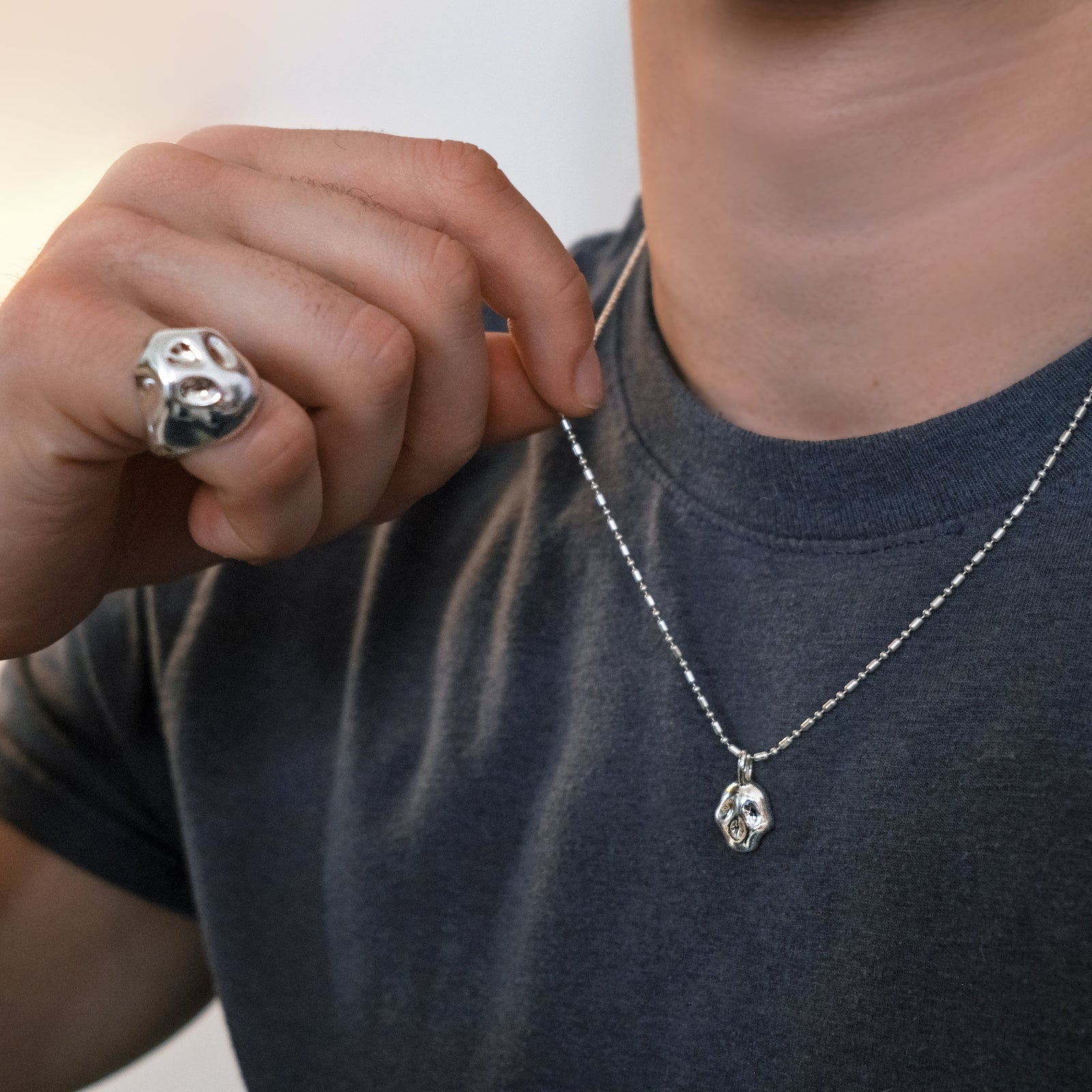 Model wearing an AMANDA PEARL recycled sterling silver molten skull ring on his pinky and a molten skull charm on a cylinder chain over a dark grey tee.
