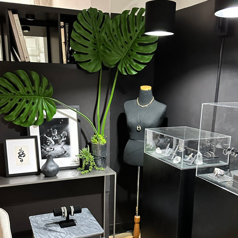 A styled jewelry display at the AMANDA PEARL showroom, with a mannequin torso wearing a gold necklace, display cases, framed art, and large green monstera leaves against a dark wall.